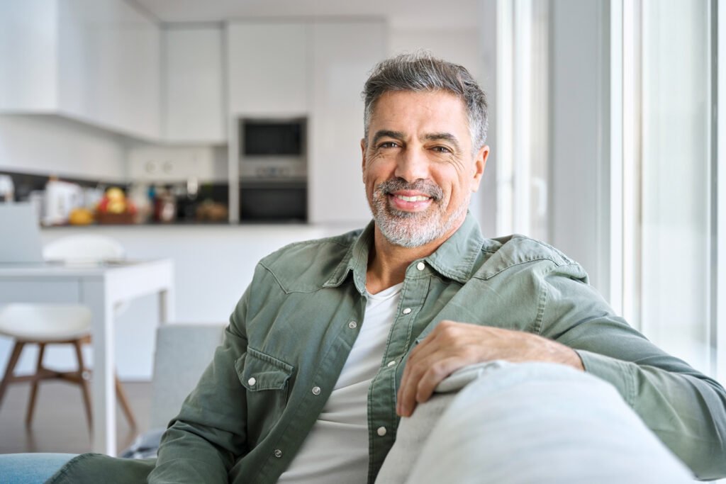 A middle-aged man with gray hair and a beard smiling while sitting on a couch in a bright modern living room.