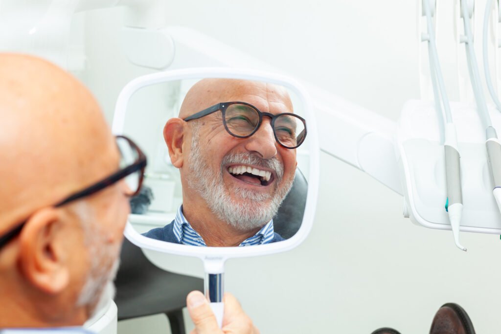 A smiling older man with glasses and a gray beard looks at his reflection in a hand mirror, checking his teeth in a dentist's office.