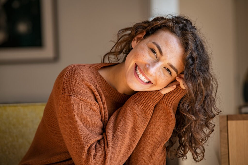 A woman with curly brown hair wearing a rust-colored sweater smiles and rests her head on her hands while seated indoors.
