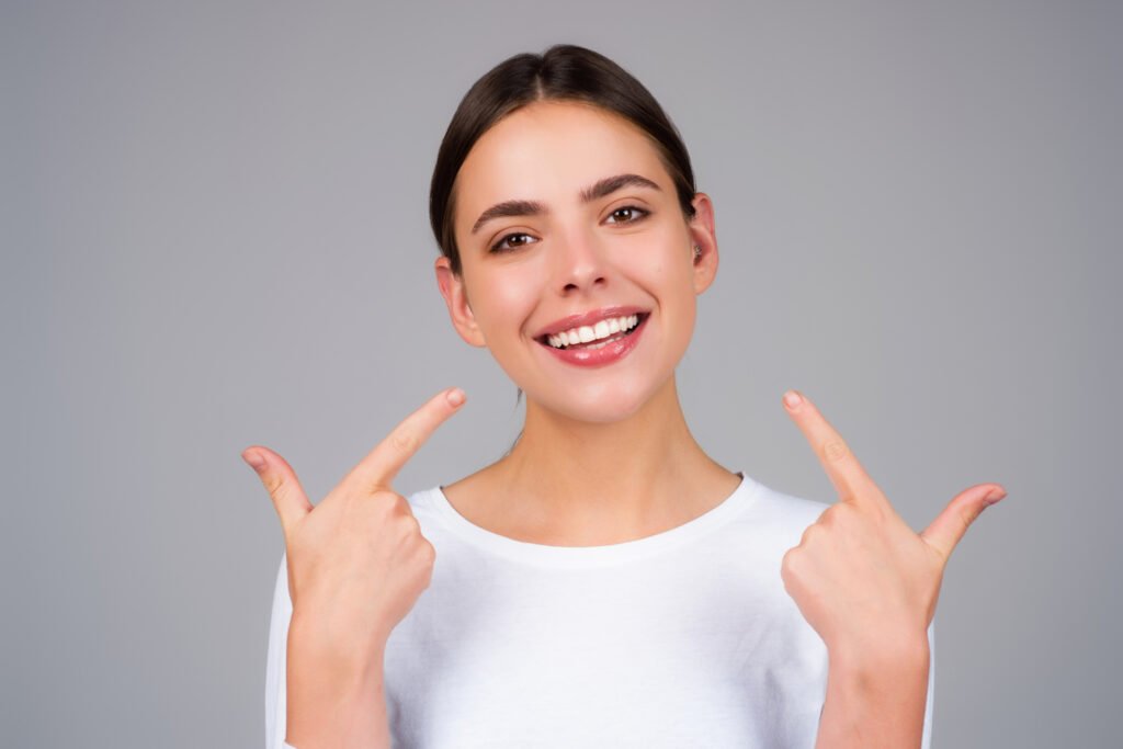 A smiling young woman with brown hair pulled back in a white shirt points to her white teeth with both index fingers, set against a gray background.
