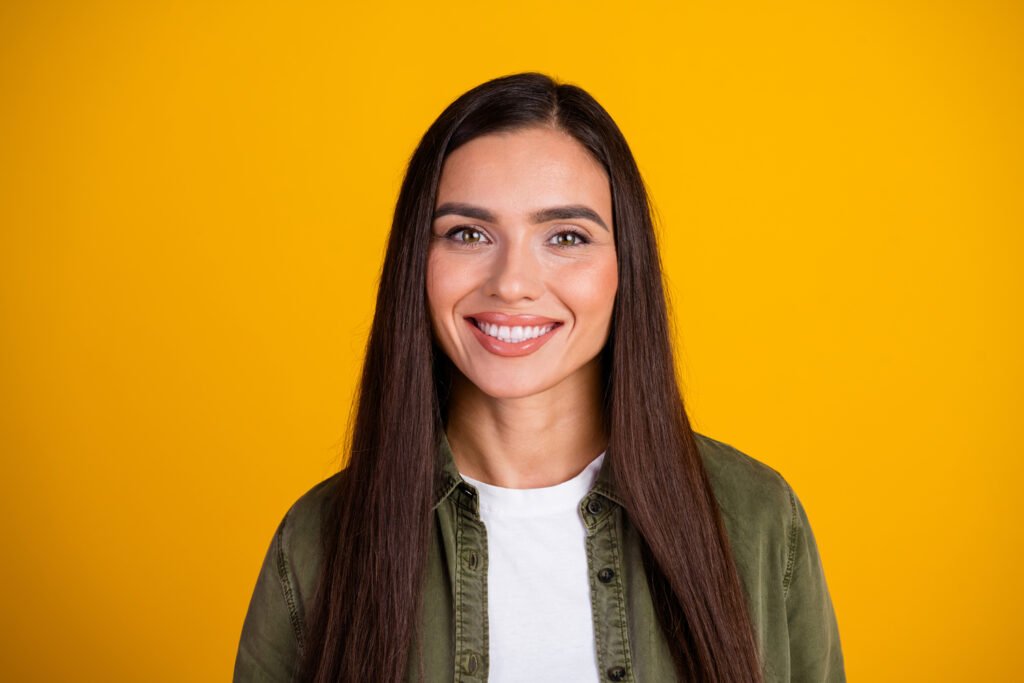 A smiling woman with long, straight dark hair and light-colored eyes, wearing a white t-shirt and an olive-green overshirt, looks at the camera against a solid yellow background.