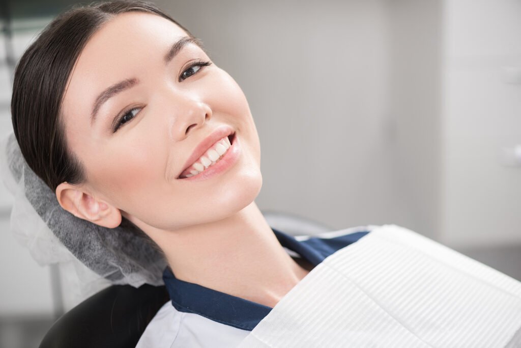 A young woman smiles brightly while reclining in a dental chair, wearing a white paper bib.