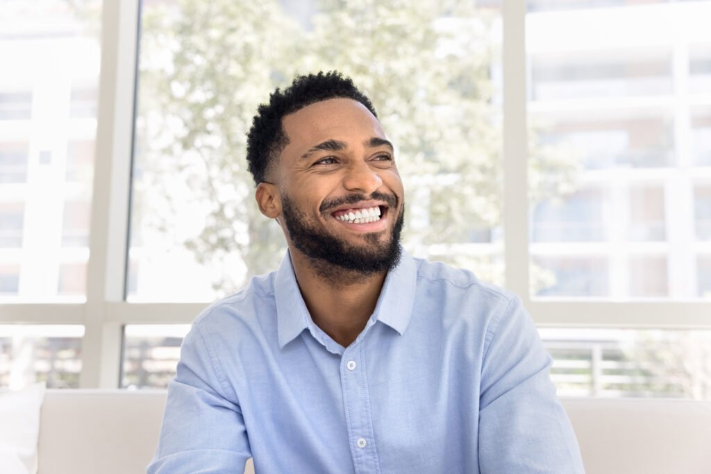 A happy man with a short beard and dark hair, wearing a light blue button-down shirt, smiles while looking off to the side. He is sitting indoors in front of a large window with bright natural light