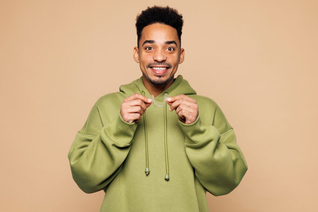 A smiling young man in a green hoodie holding a clear orthodontic aligner with both hands against a beige background.